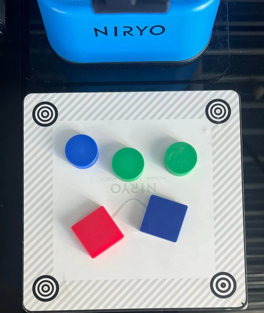 Vue de dessus d’un plan de travail portant deux disques verts, un disque bleu, un carré rouge et un carré bleu, entouré de quatre repères de calibration concentriques.