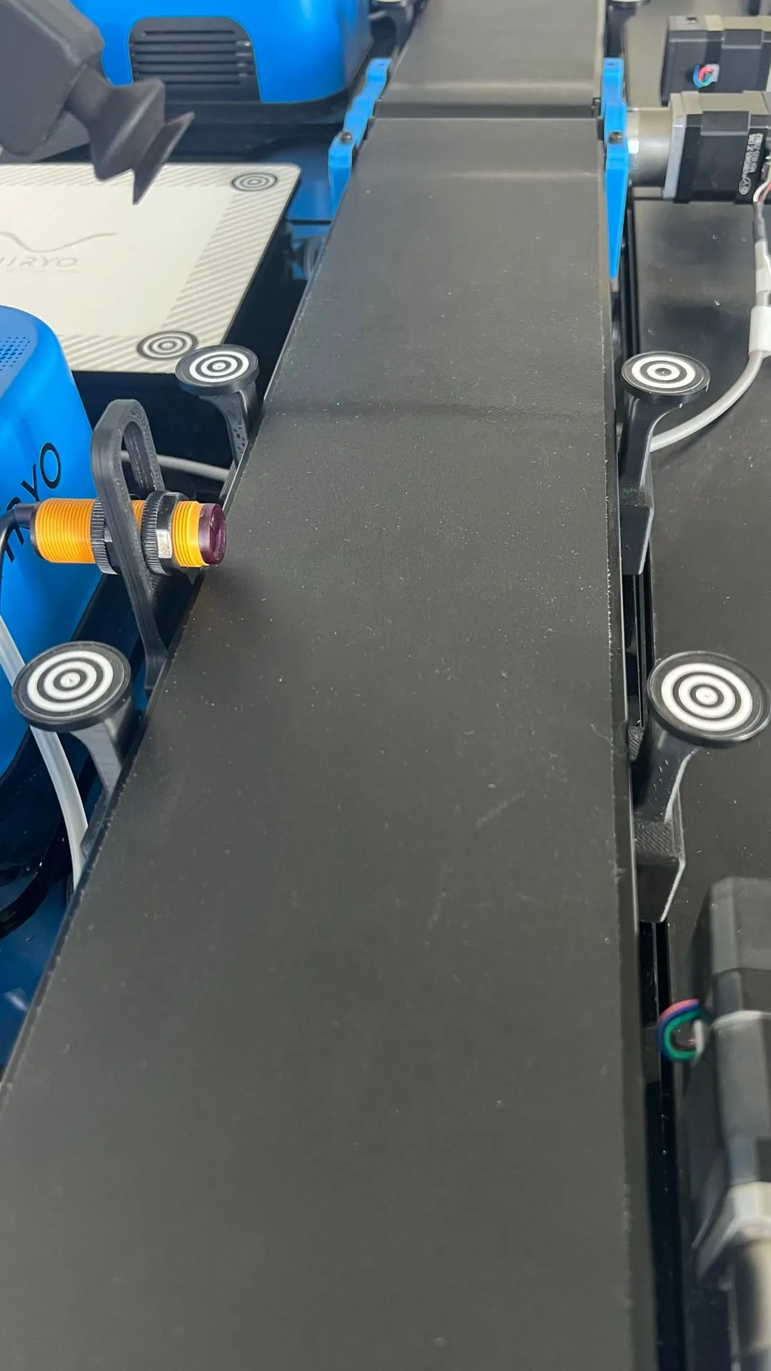 Gros plan sur un convoyeur noir, avec un capteur optique jaune fixé sur le bord et des repères de calibration.